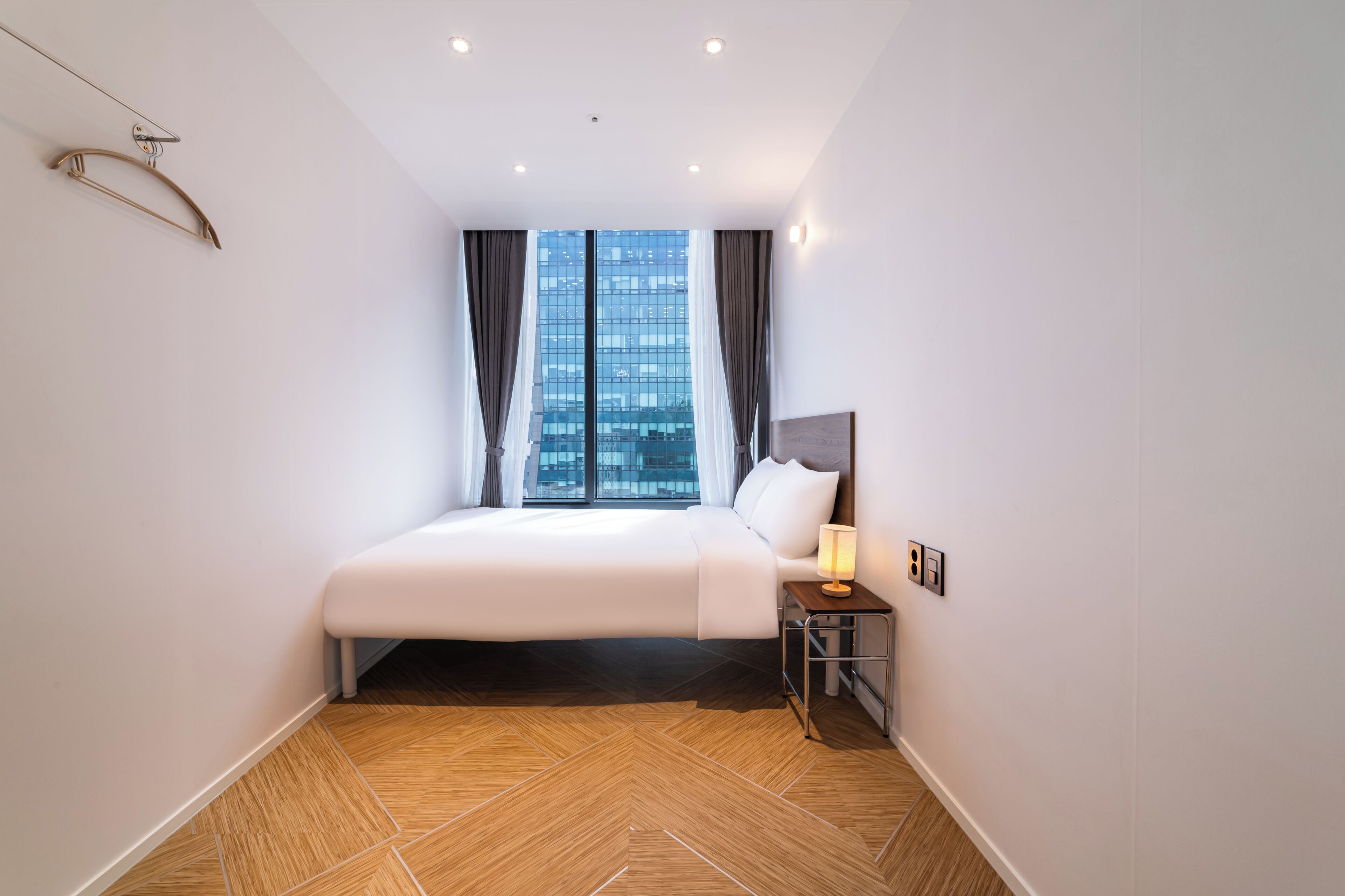 Ein modernes Hotelzimmer mit einem großen Bett, einem Nachttisch mit Lampe, Blick auf die Stadt durch große Fenster und einem Parkettboden mit Fischgrätmuster.