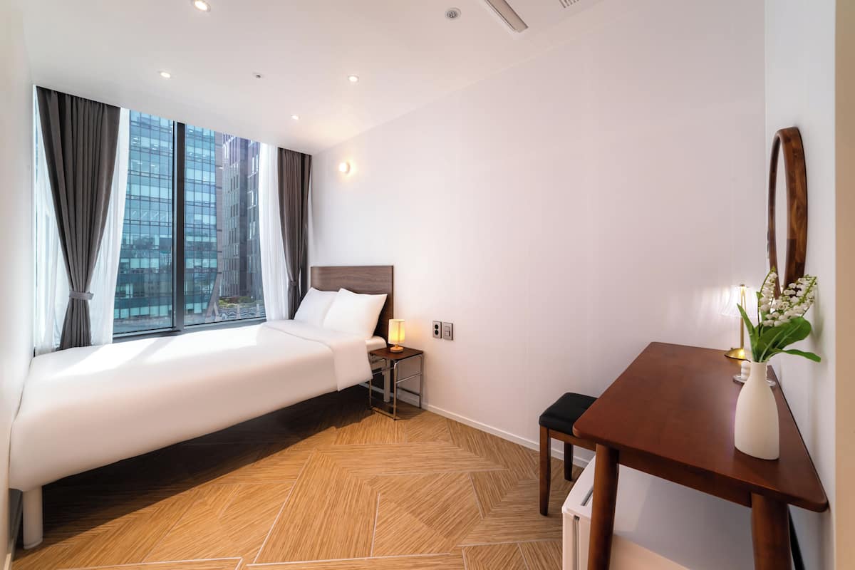 Una habitación de hotel moderna con una cama grande, un escritorio, una silla, un florero con flores y vistas a un paisaje urbano a través de grandes ventanales.