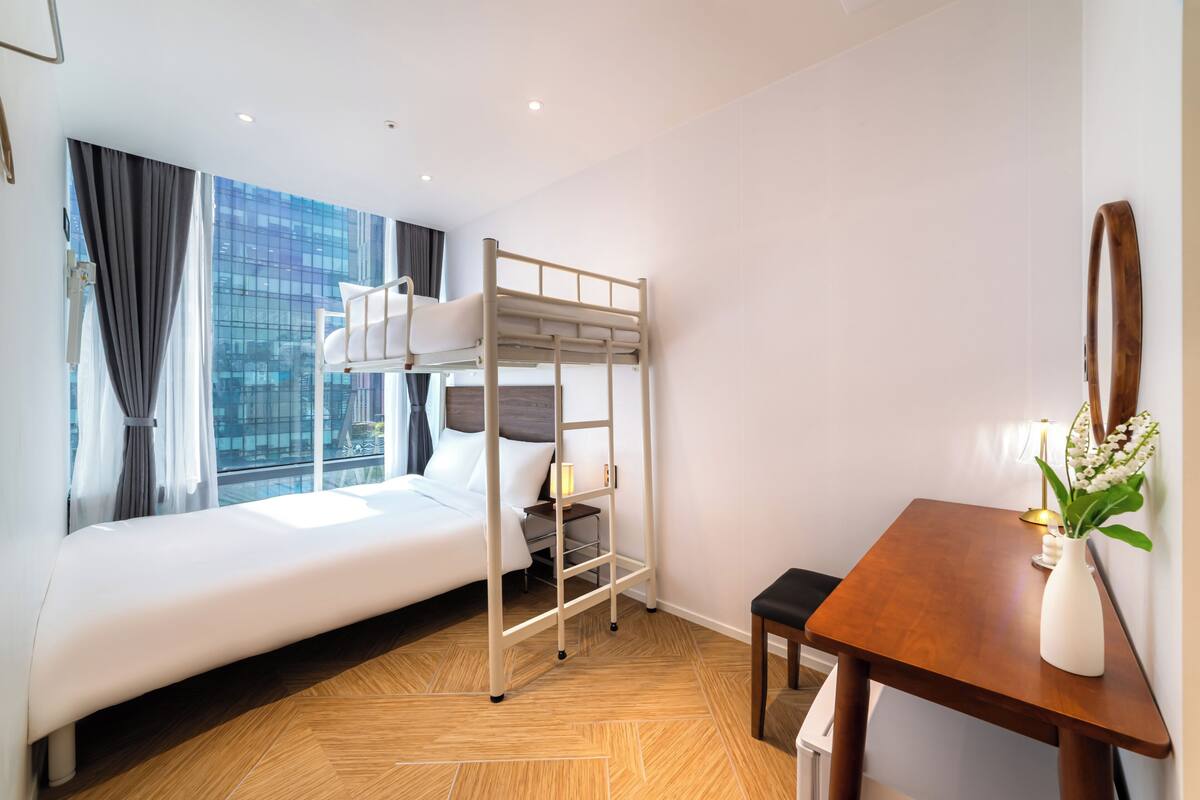 Habitación de hotel con literas, una mesita de noche con un florero y vistas a la ciudad a través de amplias ventanas.