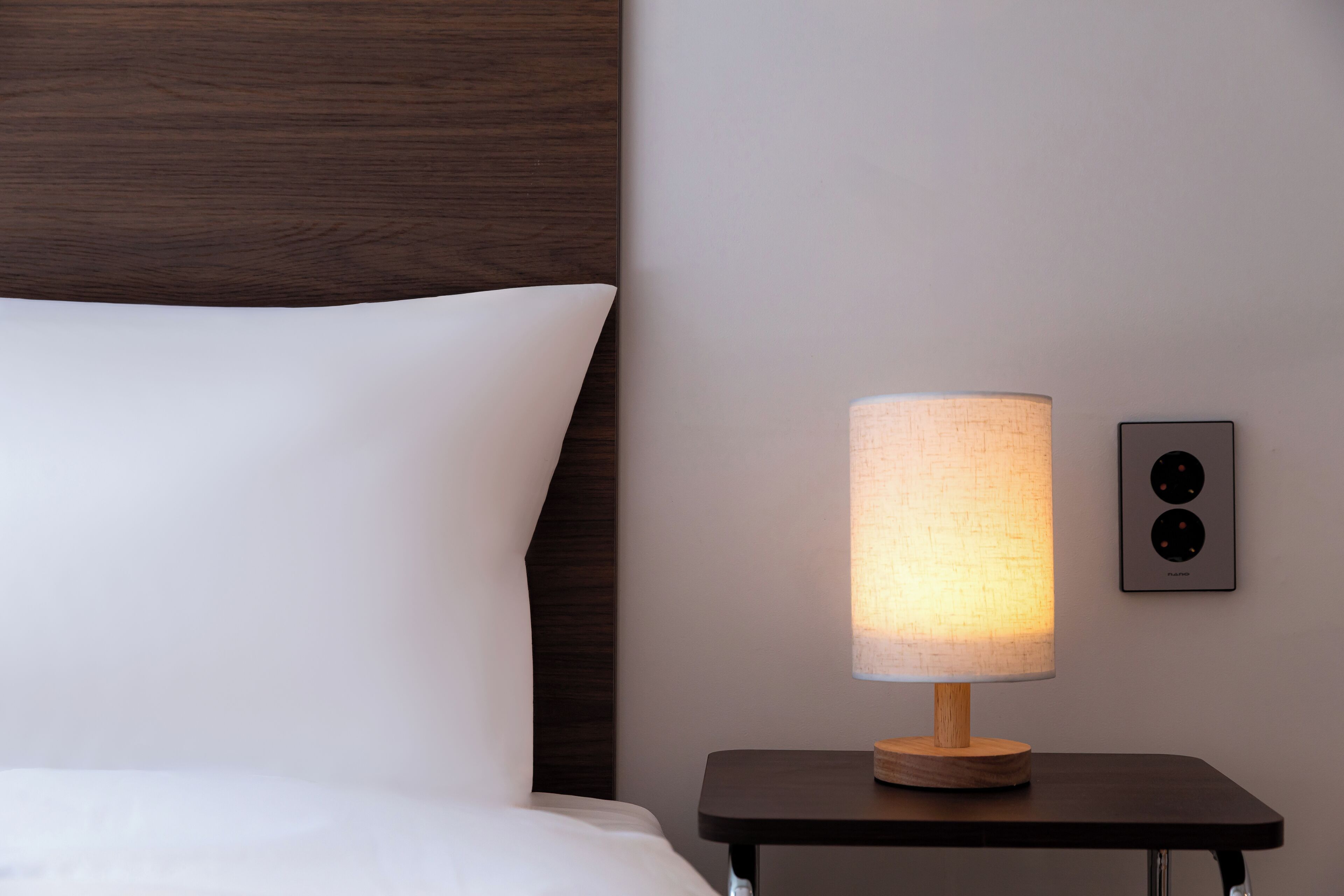Una cama bien hecha con ropa de cama blanca, una mesita de noche con una lámpara y una lámpara de pared.