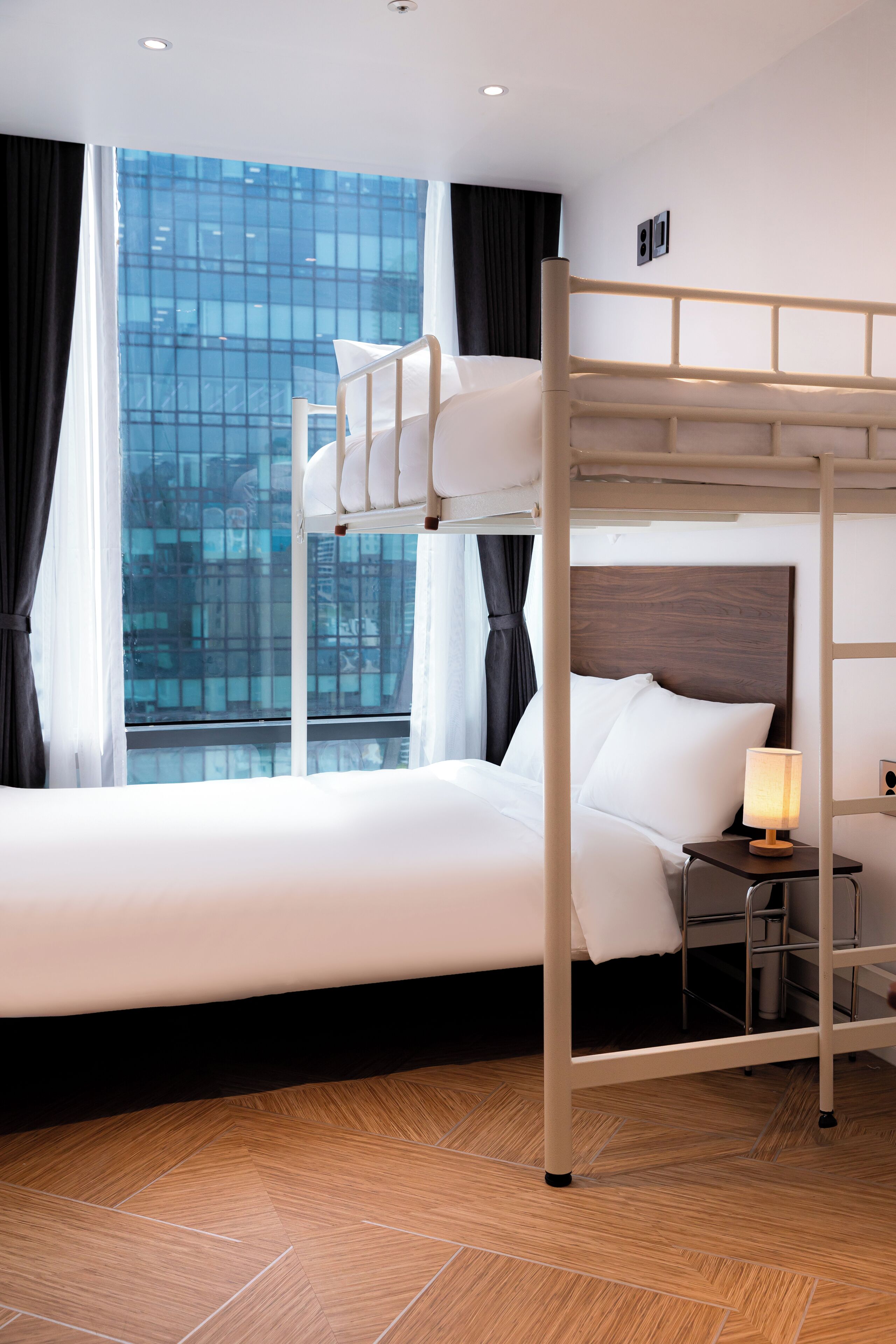 Une chambre d’hôtel avec un lit superposé, une table de chevet avec une lampe et une grande fenêtre avec des rideaux.