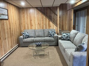A living room with a grey sofa set, a glass coffee table, and wooden paneling on the walls. - Spacious, rustic 4 bedroom cottage ideal for families, close to the beach. (Port Franks)
