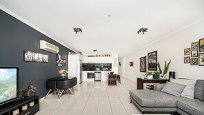 A modern living room with a grey sofa, a black coffee table, and a TV. There is a kitchen in the background with white cabinets and a dining area with a black table and chairs. - Light-filled, spacious 2 bdrm Prahran apartment with free parking. (Prahran)