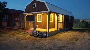 A small, single-story wooden cabin with a porch, a metal roof, and a gravel ground.