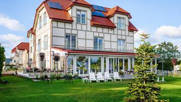 A large two-story building with a red-tiled roof, multiple windows, and a covered outdoor area with chairs and umbrellas.
