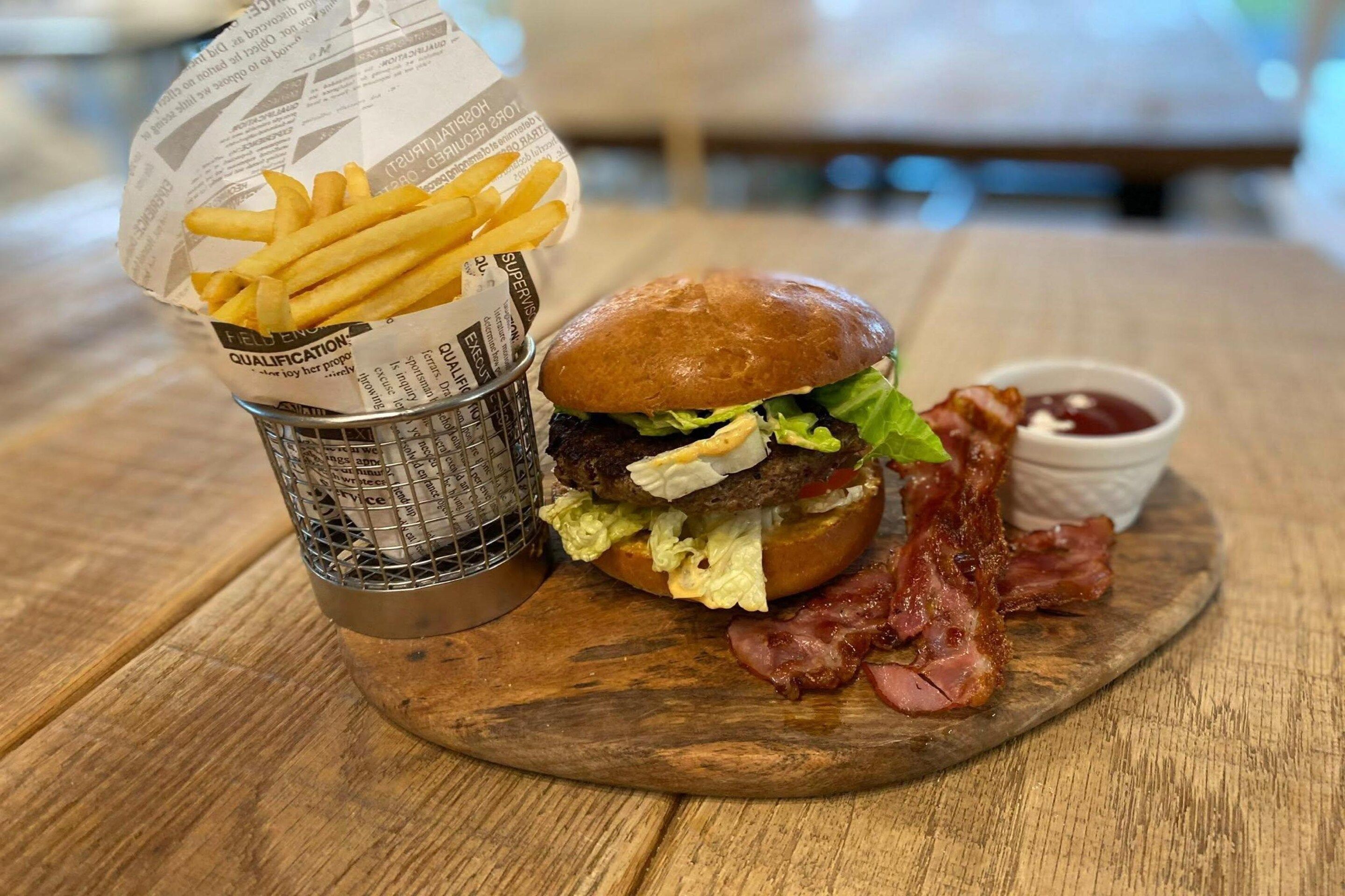 Eine Mahlzeit bestehend aus einem Burger mit Salat, Speck und einem Brötchen, begleitet von Pommes und einer kleinen Ketchup-Tasse.