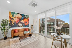 A modern living room with a large window, a sofa, a coffee table, and a dining area with a vase of flowers. - Elegant 1BD Sleeps 2 Near Independence Hall Philly (Philadelphia)