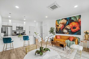 Una cocina moderna con electrodomésticos de acero inoxidable, un comedor con una mesa de mármol y una sala de estar con un sofá y un cuadro floral grande.