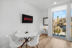 Ein modernes Zimmer mit einem weißen Tisch, weißen Stühlen, einem an der Wand montierten Fernseher, der 'NETFLIX' zeigt, und einem großen Fenster mit Blick auf Gebäude.