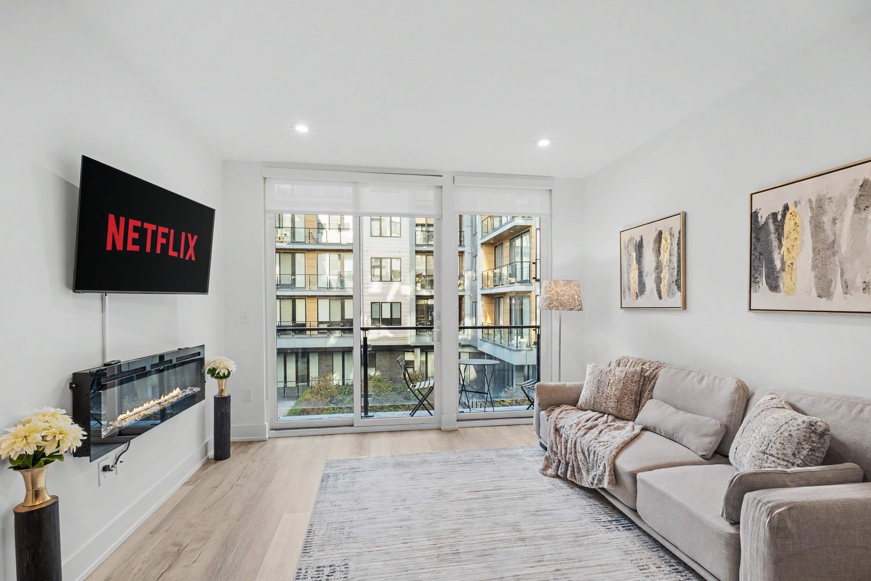 A modern living room with a large TV, a sofa, a fireplace, and a balcony with a view of buildings.