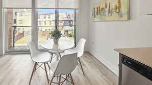 Un comedor con una mesa redonda y cuatro sillas, una ventana con vistas a edificios, y un cuadro en la pared.