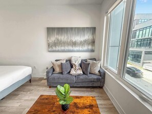 A modern living room with a grey sofa, a large abstract painting, a potted plant, and a view of the street through a large window. - Charming Studio Sleeps 2 | Walk to Liberty Bell (Philadelphia)