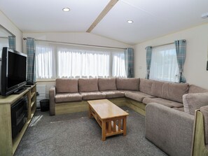 A living room with a sectional sofa, a wooden coffee table, a television on a stand, and large windows with curtains. - Caravan 325 (Lowestoft)