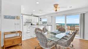 A modern kitchen with white cabinets, a dining area with a glass table and chairs, and a balcony with a view of the ocean.