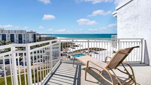 A balcony with a view of a beach and ocean, a pool, and a chair.