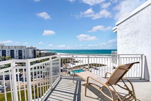 A balcony with a view of a beach and ocean, a pool, and a chair.