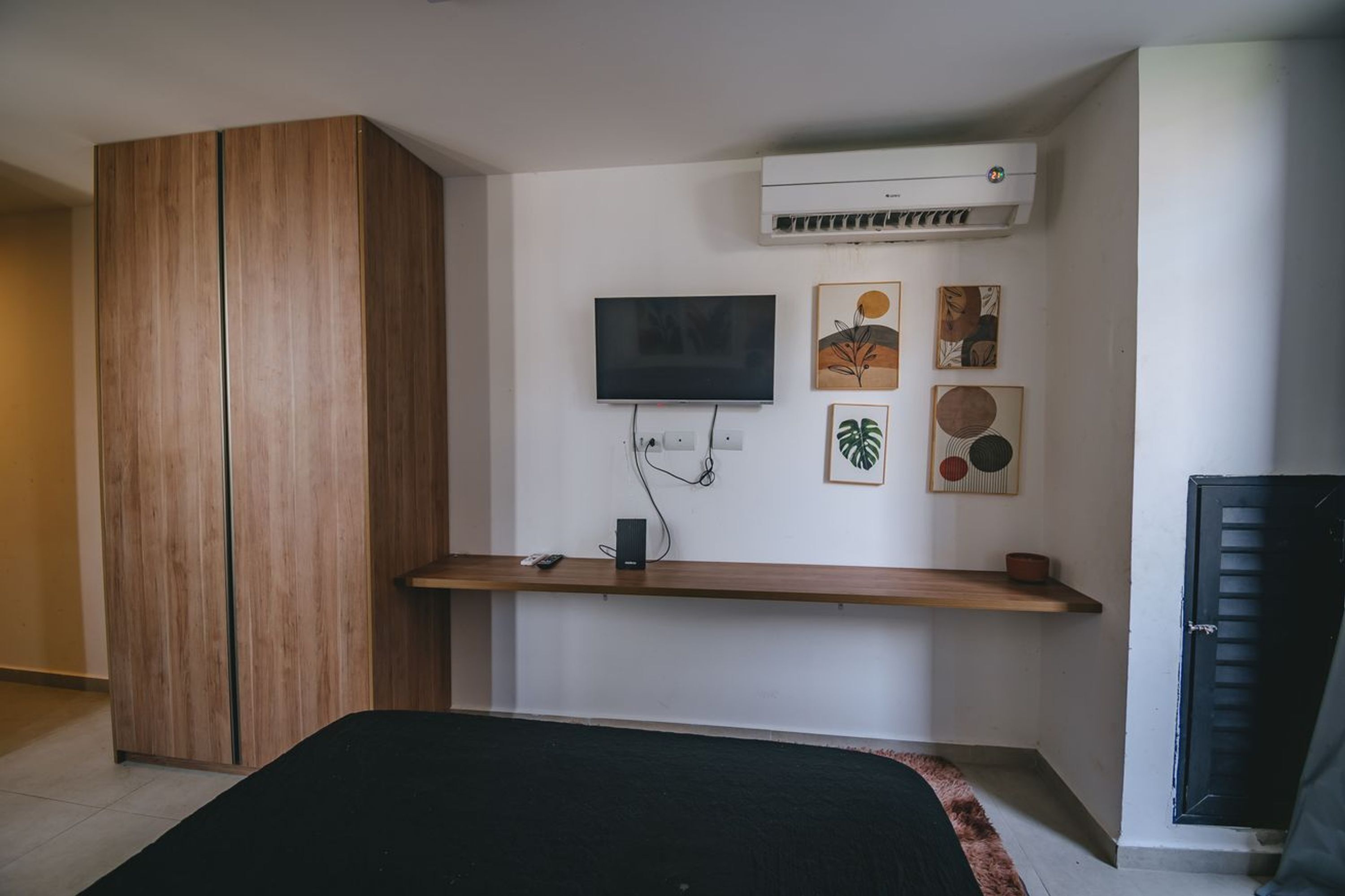 A bedroom with a bed, a TV mounted on the wall, a wooden wardrobe, and a small shelf with a remote and a cup.