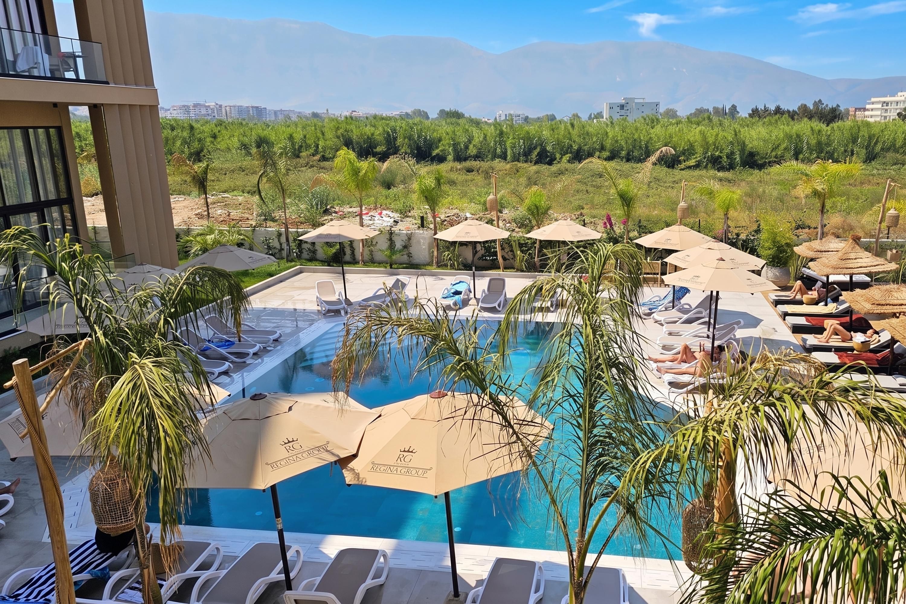 A pool area with lounge chairs and umbrellas, surrounded by greenery and a view of distant buildings.