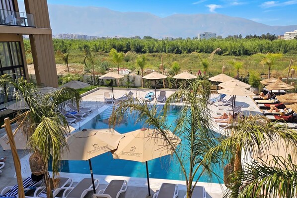 A pool area with lounge chairs and umbrellas, surrounded by greenery and a view of distant buildings.