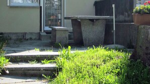 Een betonnen zitgedeelte buiten, met een trap die naar een deur leidt, omringd door groen.