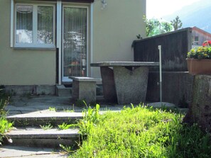 Ein betonierter Außenbereich mit Sitzgelegenheiten und Stufen, die zu einer Tür führen, umgeben von Grünpflanzen.