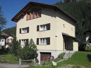 Ein zweigeschossiges Gebäude mit geneigtem Dach, zahlreichen Fenstern mit Fensterläden und einem Hauseingang mit Stufen.
