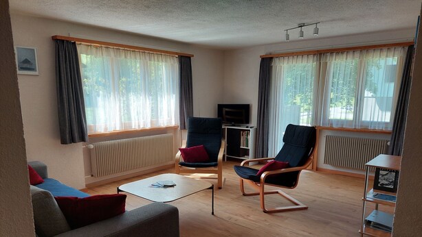 Een woonkamer met een bank, twee stoelen, een salontafel, een televisie en een boekenkast.