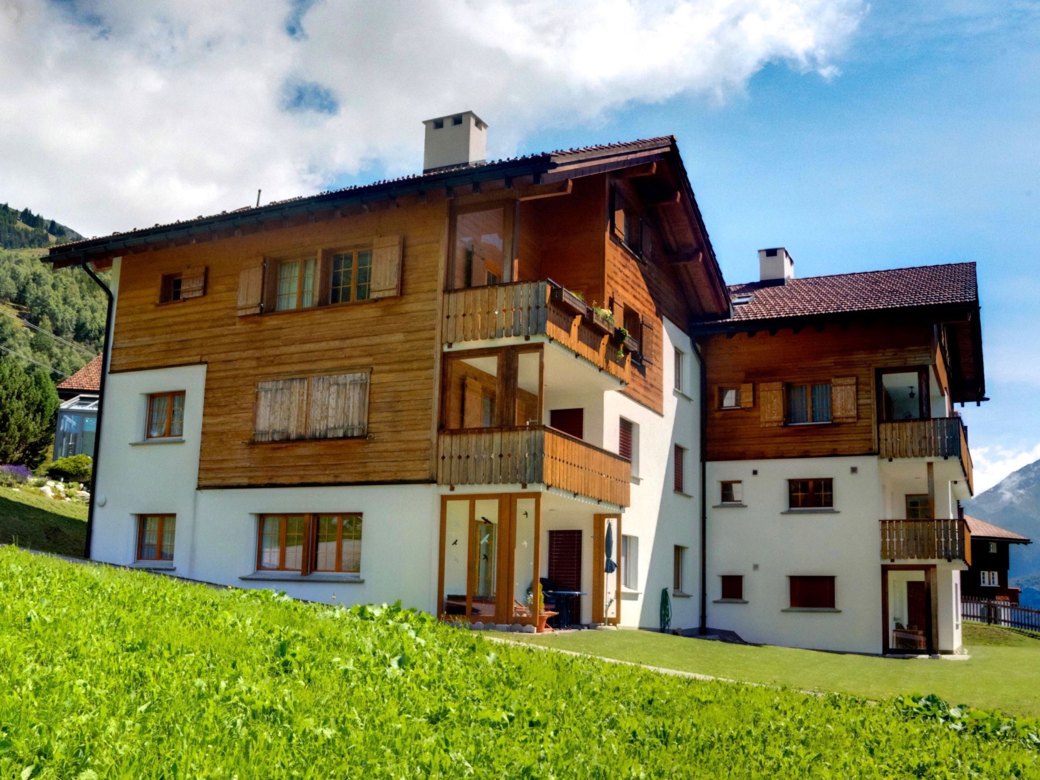 Ein zweistöckiges Holzhaus mit Balkonen, heller Fassade und einer Bergkulisse.