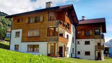 Ein zweistöckiges Holzhaus mit Balkonen, heller Fassade und einer Bergkulisse.