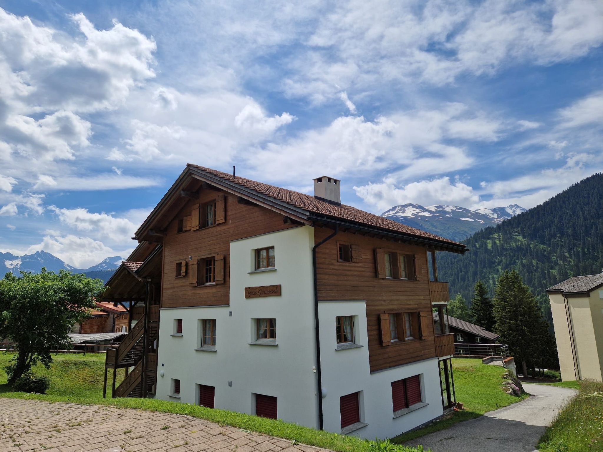 Ein zweistöckiges Holzhaus mit weißer Untergeschosse und roten Fensterläden, vor einer Bergkulisse und einem teilweise bewölkten Himmel.
