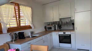 Eine kleine Küche mit weißen Schränken, einer Mikrowelle, einem Backofen und einem Waschbecken. Ein Fenster mit Vorhängen, ein Holztisch mit einem Stuhl und ein Topf mit rosa Blüten.