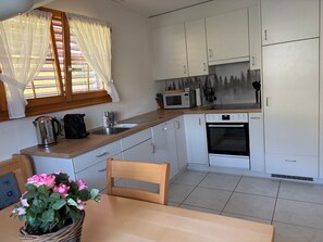 A compact kitchen with white cabinets, a microwave, an oven, and a sink. There is a window with curtains, a wooden table with a chair, and a potted plant with pink flowers. - Tgèsa Garnetsch No. 1 Young, (Sedrun), 170.01 by Interhome (Sedrun)