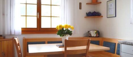 A dining area with a wooden table and chairs, a vase with yellow flowers, and a window with white curtains.