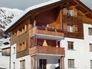Un edificio de madera de varios pisos con balcones, una pista de esquí al fondo y una montaña.