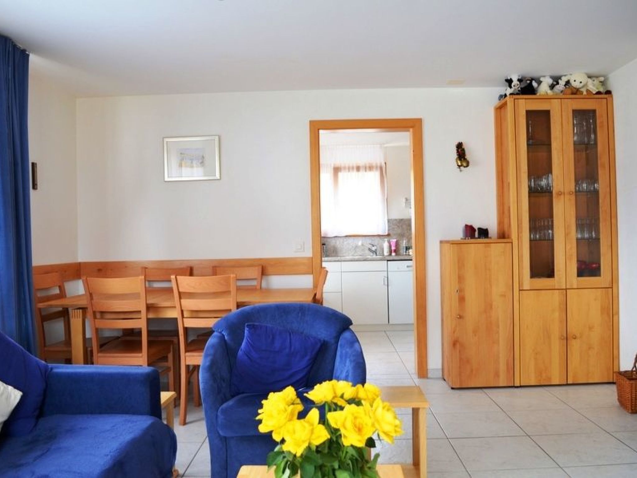 Uma sala de estar com um sofá azul, cadeiras de madeira, um armário de madeira e uma mesa pequena com flores amarelas.