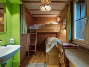 Ein kleines Zimmer mit Etagenbett, einem Holztisch, einem Fenster, einem Waschbecken und einer Treppe.