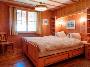 Een kamer met houten vloer, een bed, een stoel, een raam met gordijnen en een wandplank met een foto.