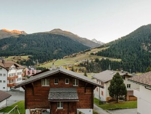 Ein Holzhaus mit geneigtem Dach, umgeben von anderen Häusern und einer gebirgigen Landschaft.