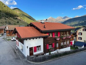 Un chalet traditionnel avec un toit recouvert de tuiles rouges, une façade en bois et des volets rouges, qui se détache sur un paysage de montagnes et d’un ciel dégagé.