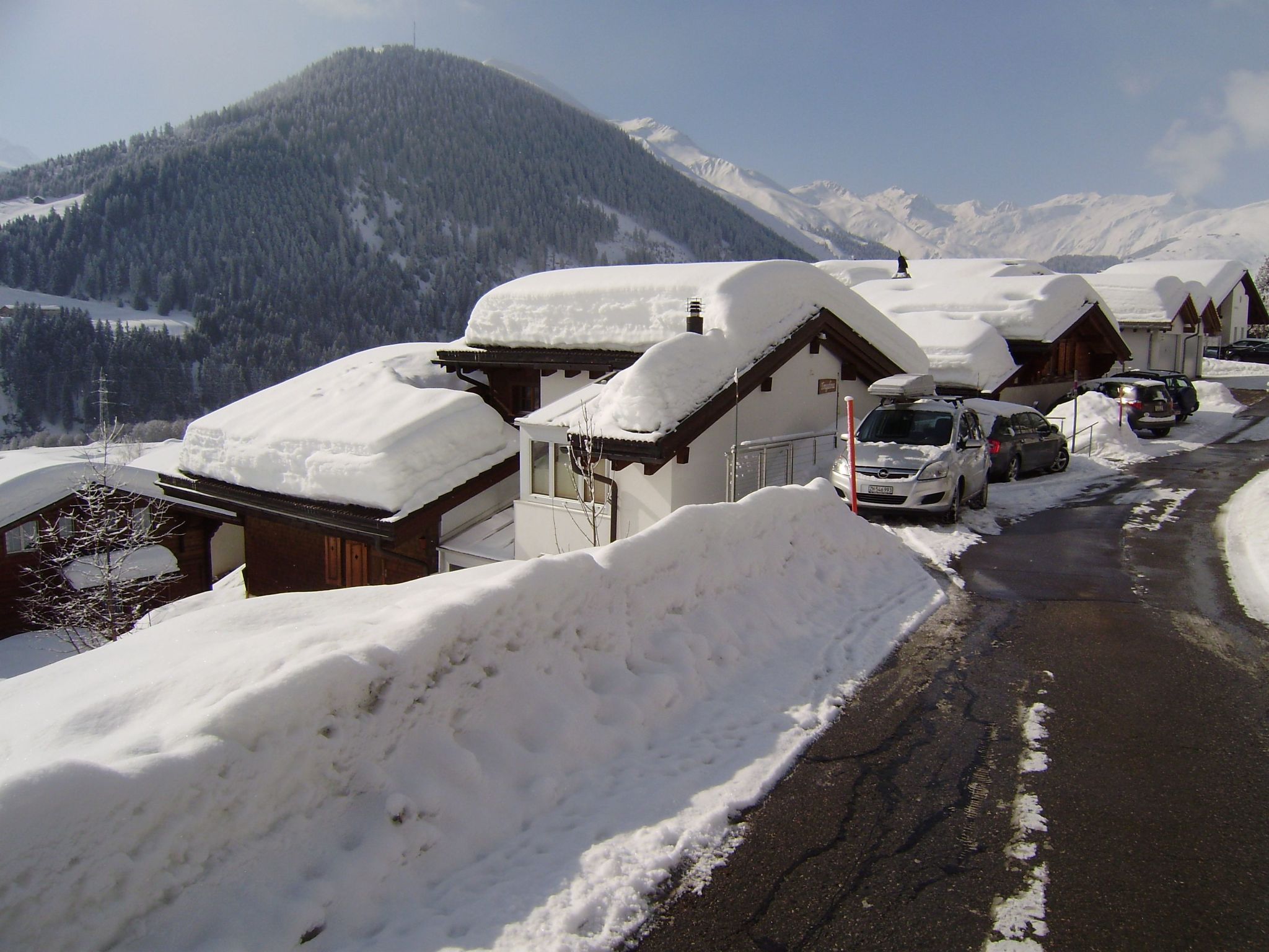 A snow-covered mountain road with wooden buildings, a parked car, and a snowbank.