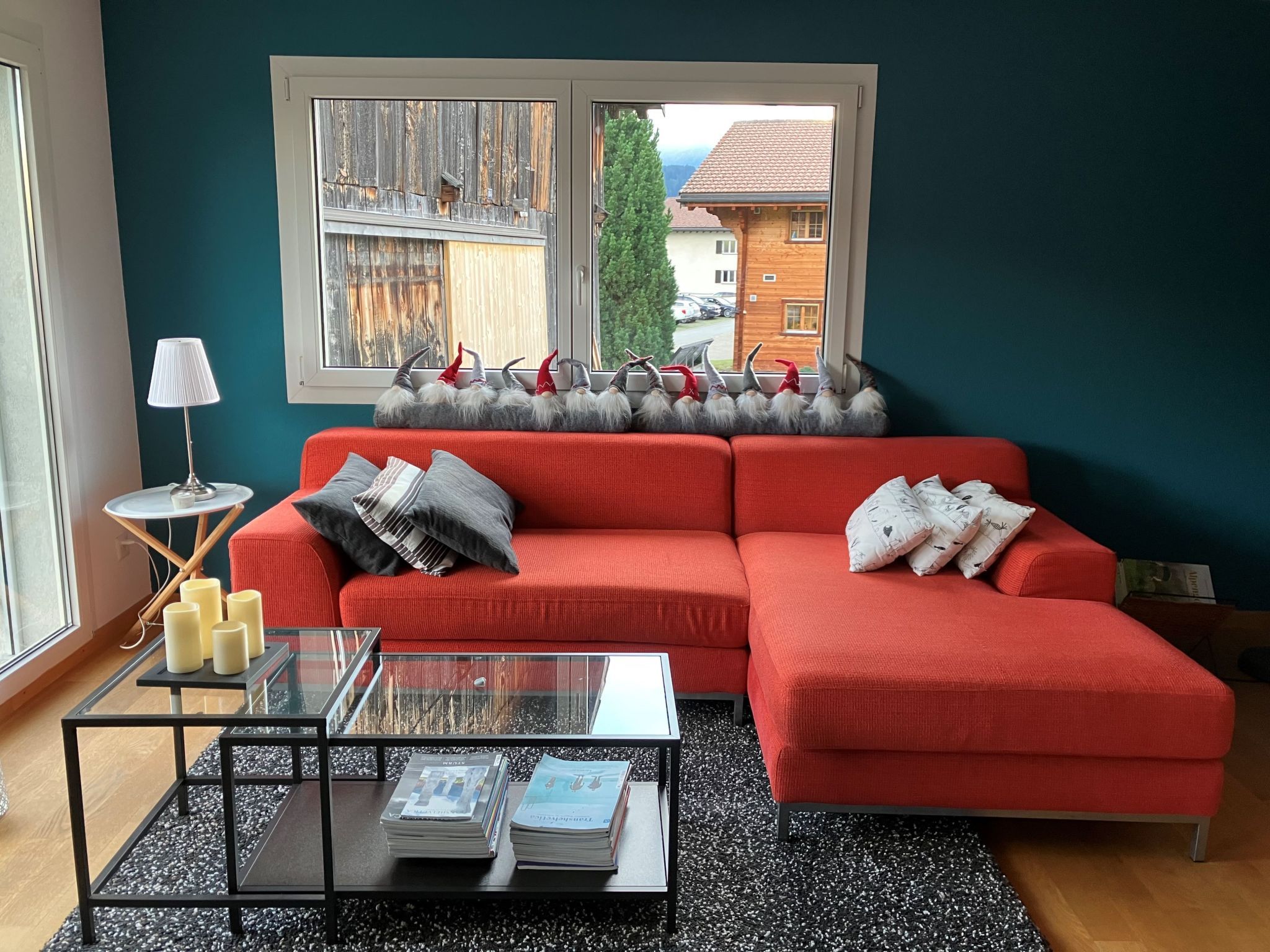 Ein modernes Wohnzimmer mit einem roten Sofa, einem Glastisch mit Zeitschriften und Blick auf ein Holzhaus durch ein Fenster.