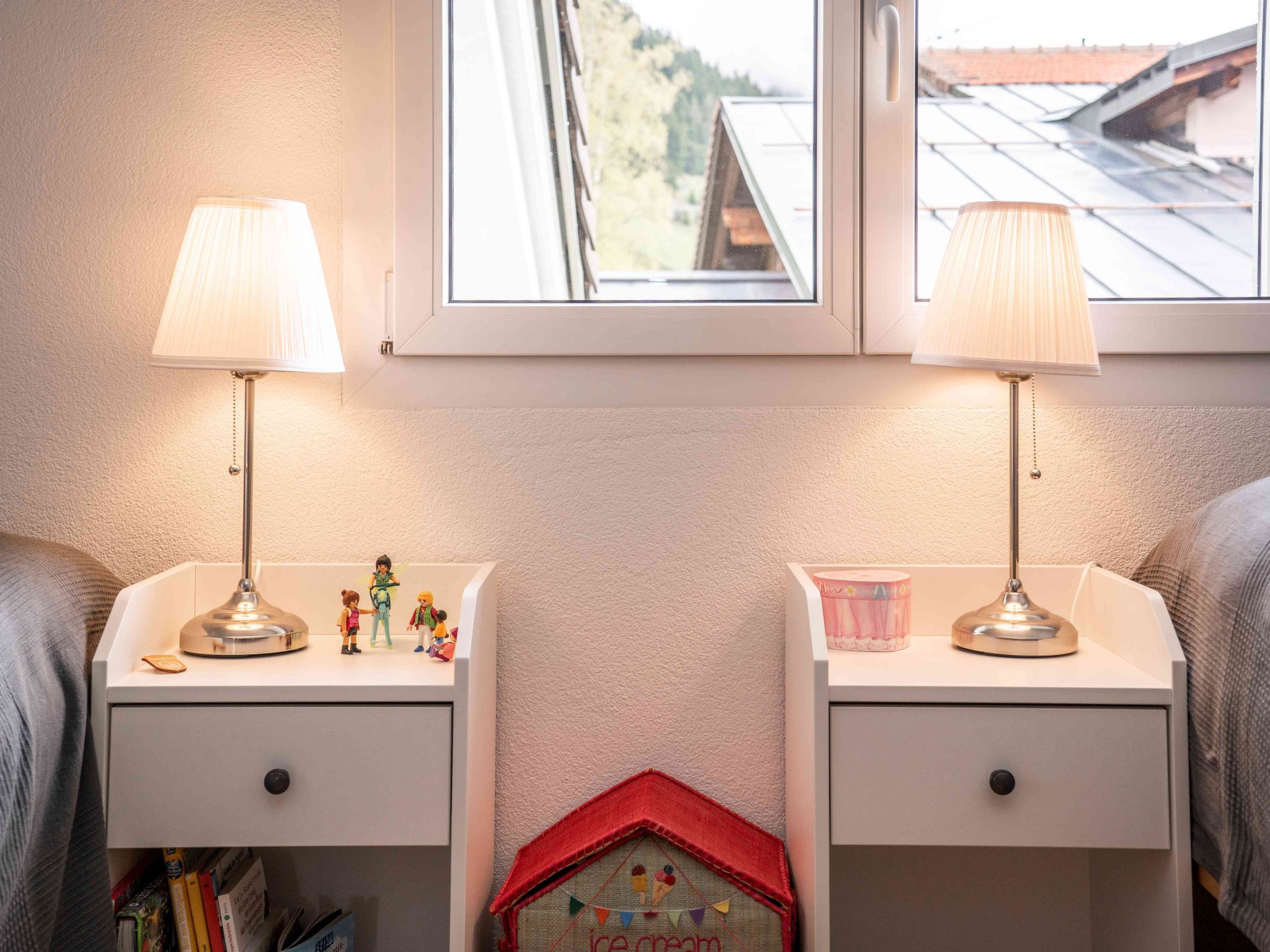 Two bedside tables with lamps, a small toy house, and a window with a view of a building.