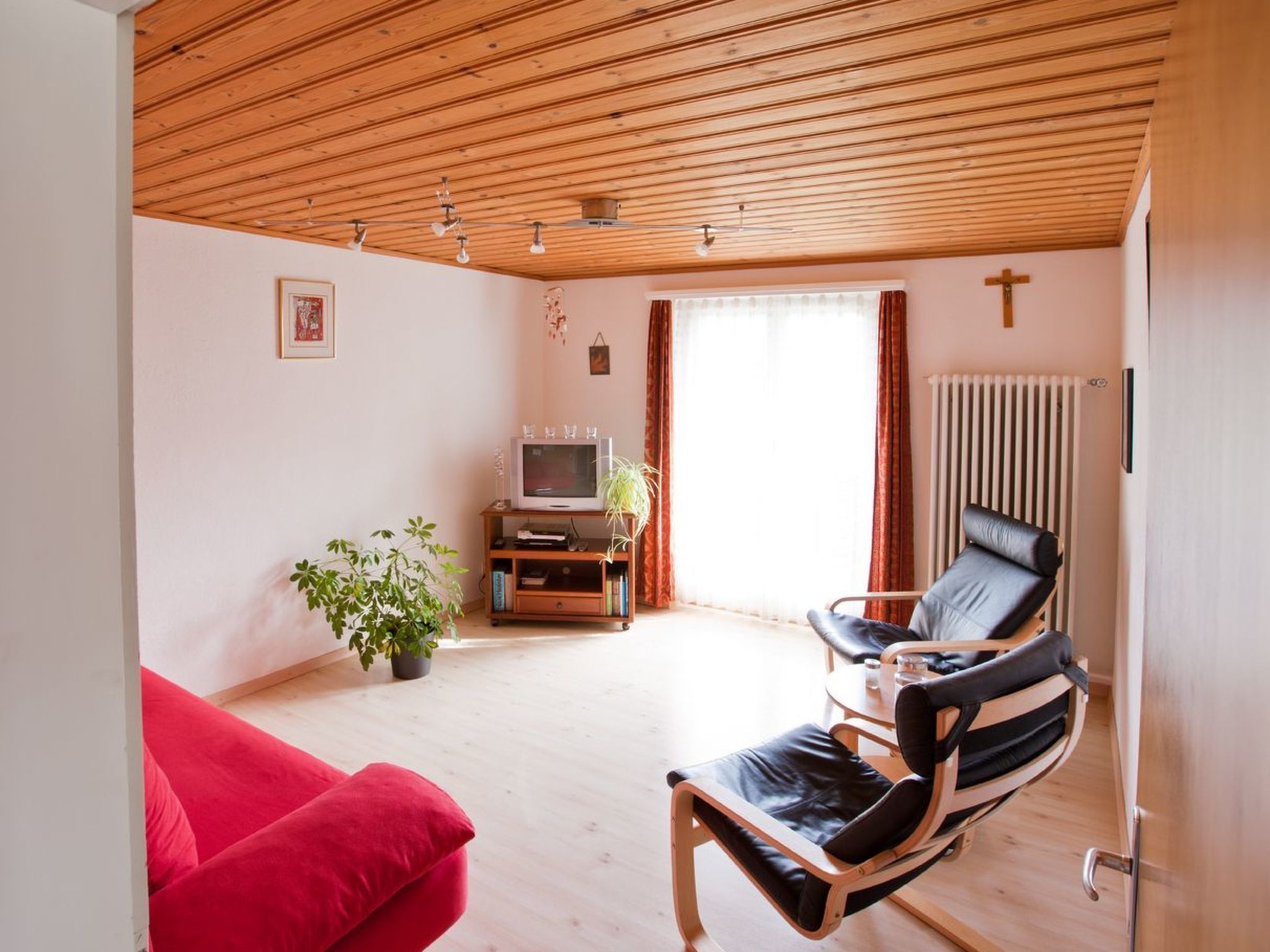 Een woonkamer met een rode bank, een zwarte lederen fauteuil, een houten plafond, een radiator en een televisie.