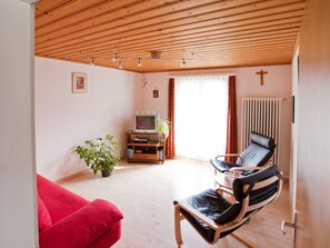 Ein Wohnzimmer mit einem roten Sofa, einem schwarzen Ledersessel, einer Holzbalkendecke, einem Heizkörper und einem Fernseher.