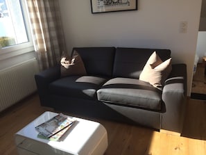 A modern living room with a sofa, two pillows, a coffee table with magazines, a window with curtains, and a framed picture on the wall. - La Sclarida Kissling, (Segnas/Disentis), 1630 by Interhome (Disentis)