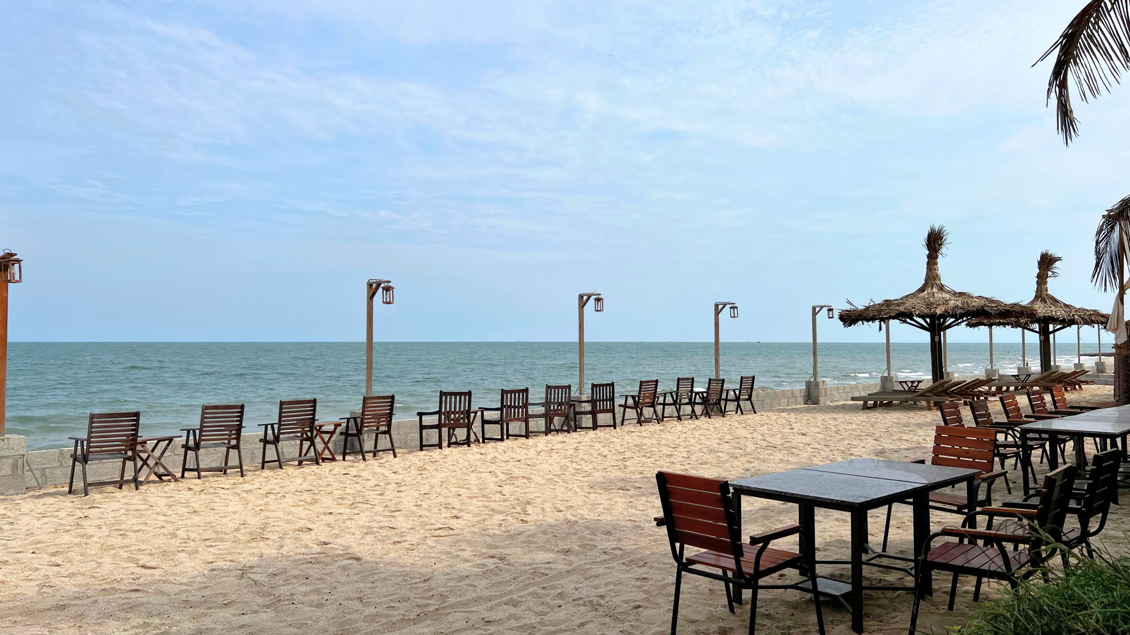 A beachside seating area with chairs and tables, a thatched-roof structure, and a view of the ocean.