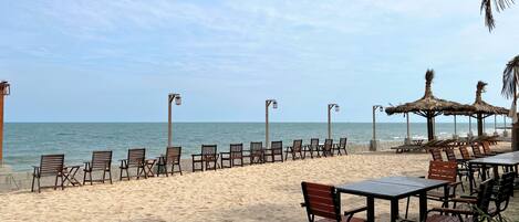 A beachside seating area with chairs and tables, a thatched-roof structure, and a view of the ocean.