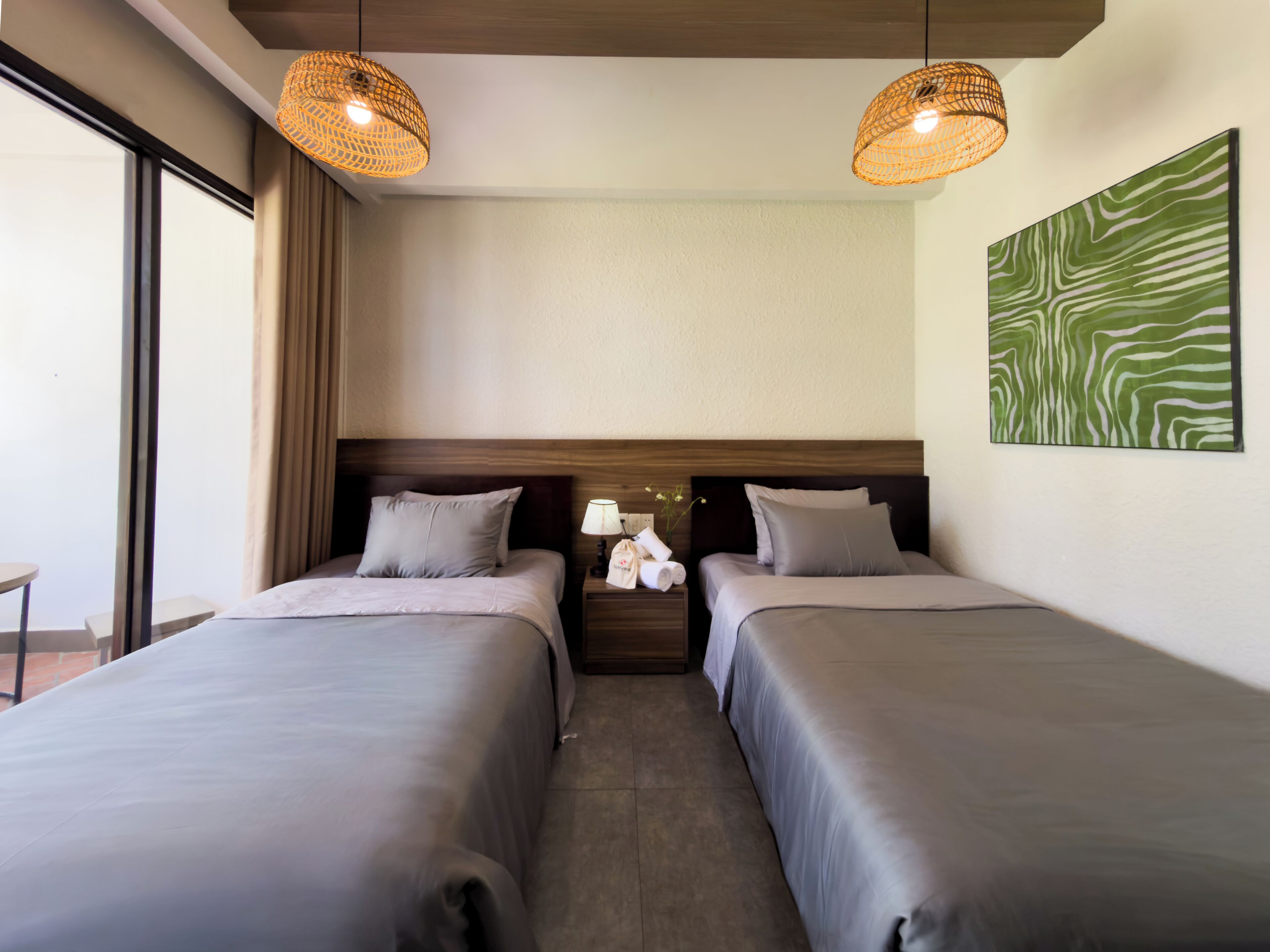 Une chambre d’hôtel avec deux lits, une table de chevet avec une lampe, un tableau décoratif au mur et une fenêtre avec des rideaux.
