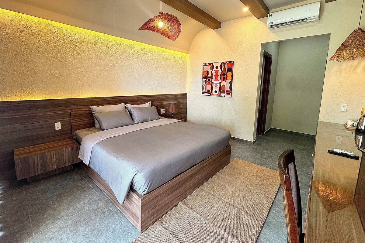 A modern bedroom with a wooden headboard, a bed with grey bedding, a woven rug, and a hanging light fixture.