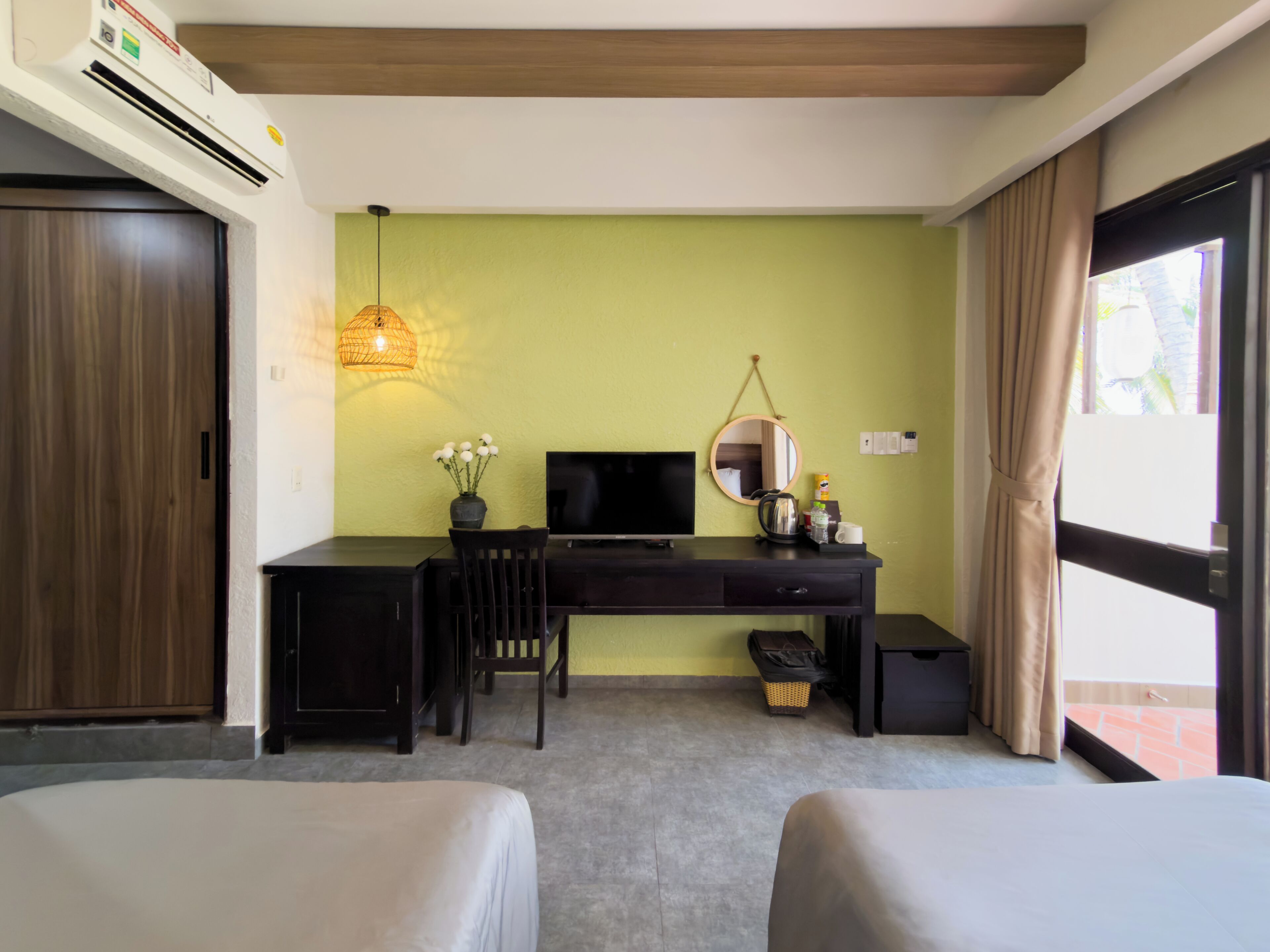 Habitación de hotel con escritorio, dos camas, televisor, espejo, lámpara colgante y ventana con cortinas.